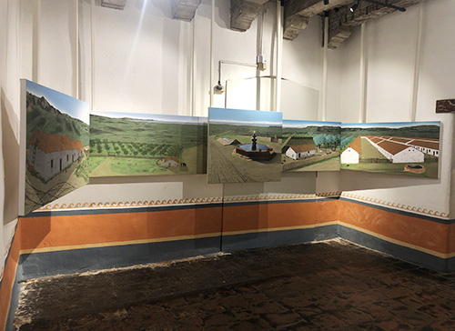 The south side of the gallery displays five images. From left to right they are the Majordomo's house, the orchard, the fountain and lavadora, the garden, and Chumash Pueblo. The images hang dimensionally from the ceiling in relation to how they would have appeared from the front of the Mission. 